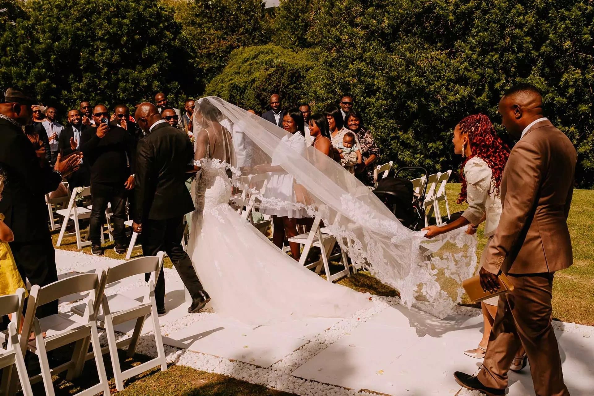A bride walking down a white aisle lined with white folding chairs while guests watch (stock photo)