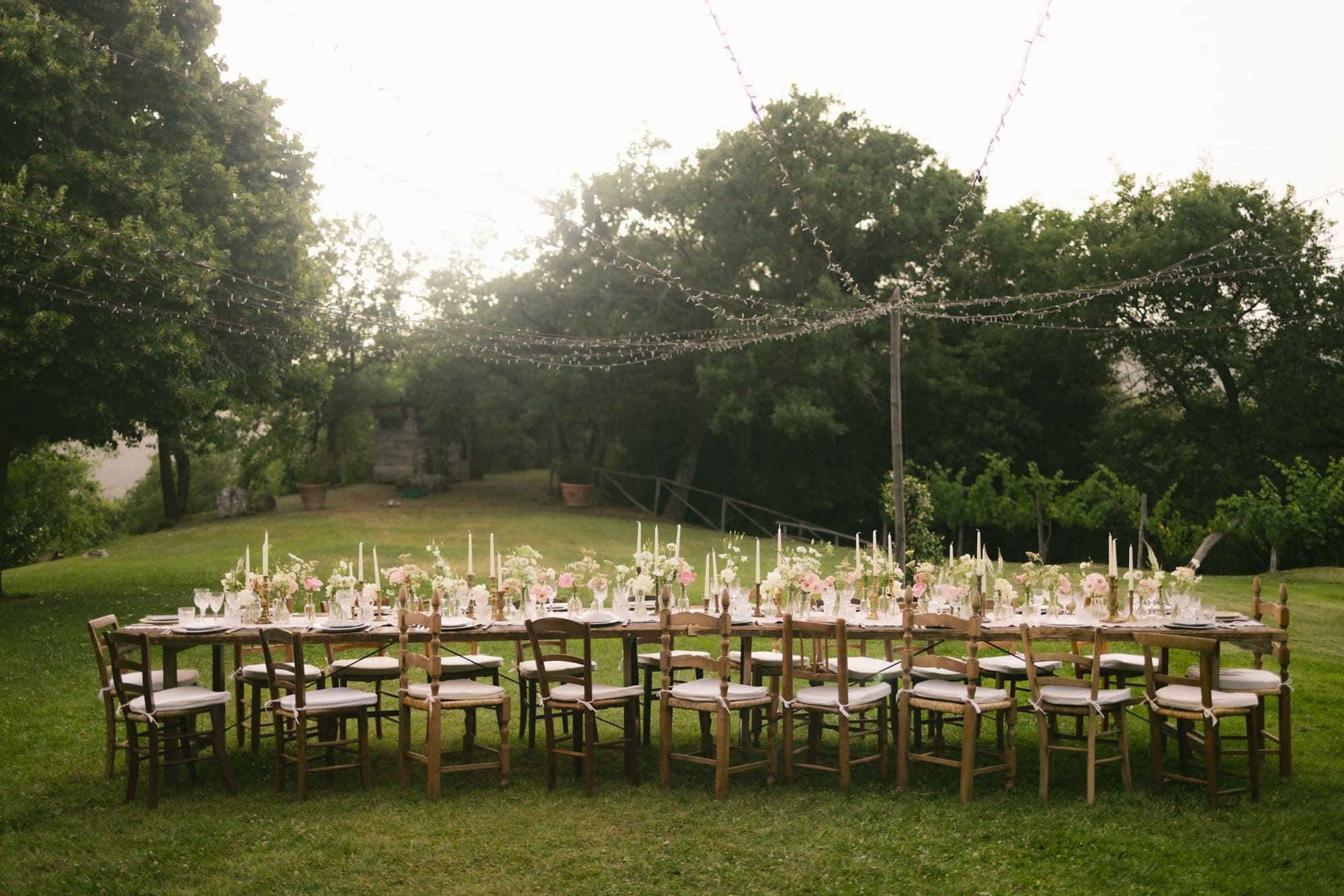 A long garden table set with flowers and candles at dusk under string lights (stock photo)